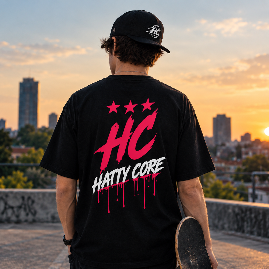 A person stands on a rooftop at sunset, wearing a black oversized t-shirt featuring a bold pink and white graphic with the text HC HATTY CORE and three stars.