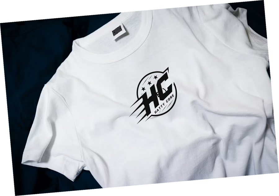 A white t-shirt is laid flat on a dark blue background, featuring a circular logo in the center. The logo includes the letters HC with stylized wings and five stars above it, giving a dynamic and modern appearance.